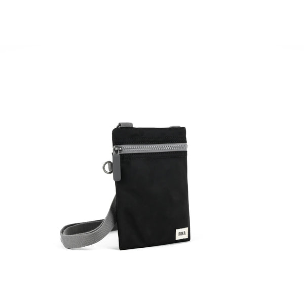 A photo of the front and side of a small rectangular black pocket bag. It has a grey zip horizontally at the top, grey straps, and a small ROKA logo in the bottom right corner. 