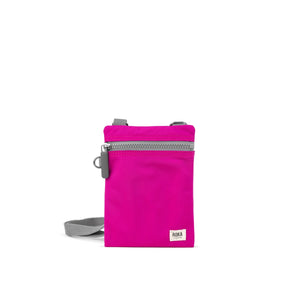 A photo of the front of a small rectangular bright pink pocket bag. It has a grey zip horizontally at the top, grey straps, and a small ROKA logo in the bottom right corner.