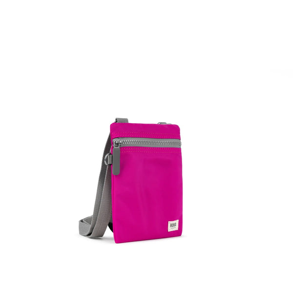 A photo of the front and side of a small rectangular bright pink pocket bag. It has a grey zip horizontally at the top, grey straps, and a small ROKA logo in the bottom right corner.