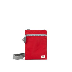 A photo of the front of a small rectangular red pocket bag. It has a grey zip horizontally at the top, grey straps, and a small ROKA logo in the bottom right corner.