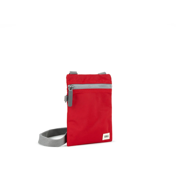A photo of the front and side of a small rectangular red pocket bag. It has a grey zip horizontally at the top, grey straps, and a small ROKA logo in the bottom right corner. 