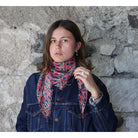 A woman wearing a colorful abstract-patterned scarf and a dark denim jacket, standing against a rustic stone wall.
