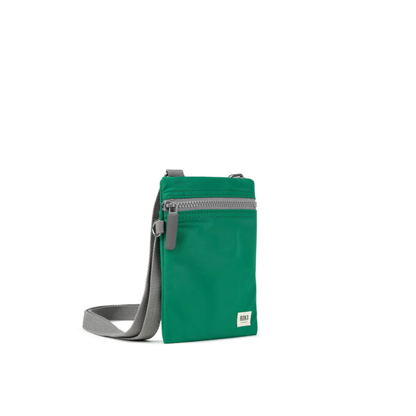 A photo of the front and side of a small rectangular emerald green pocket bag. It has a grey zip horizontally at the top, grey straps, and a small ROKA logo in the bottom right corner. 