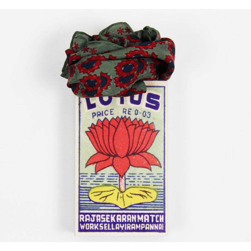 A folded fabric hair scrunchie with a red and navy floral pattern on an olive green base, displayed on top of a vintage Lotus matchbox against a plain white background.