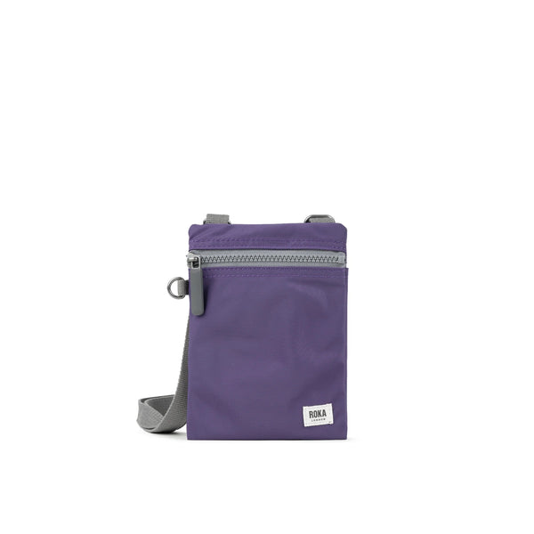 A photo of the front of a small rectangular purple pocket bag. It has a grey zip horizontally at the top, grey straps, and a small ROKA logo in the bottom right corner.