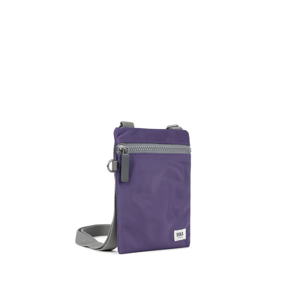 A photo of the front and side of a small rectangular purple pocket bag. It has a grey zip horizontally at the top, grey straps, and a small ROKA logo in the bottom right corner.