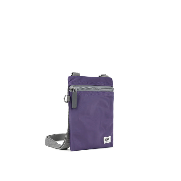 A photo of the front and side of a small rectangular purple pocket bag. It has a grey zip horizontally at the top, grey straps, and a small ROKA logo in the bottom right corner. 