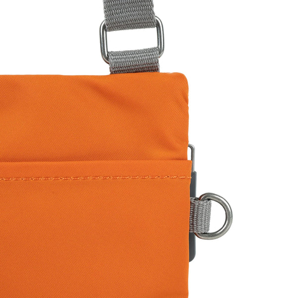 A close up photo of the back of an orange bag, showing the grey strap and neatly stitched pocket opening.