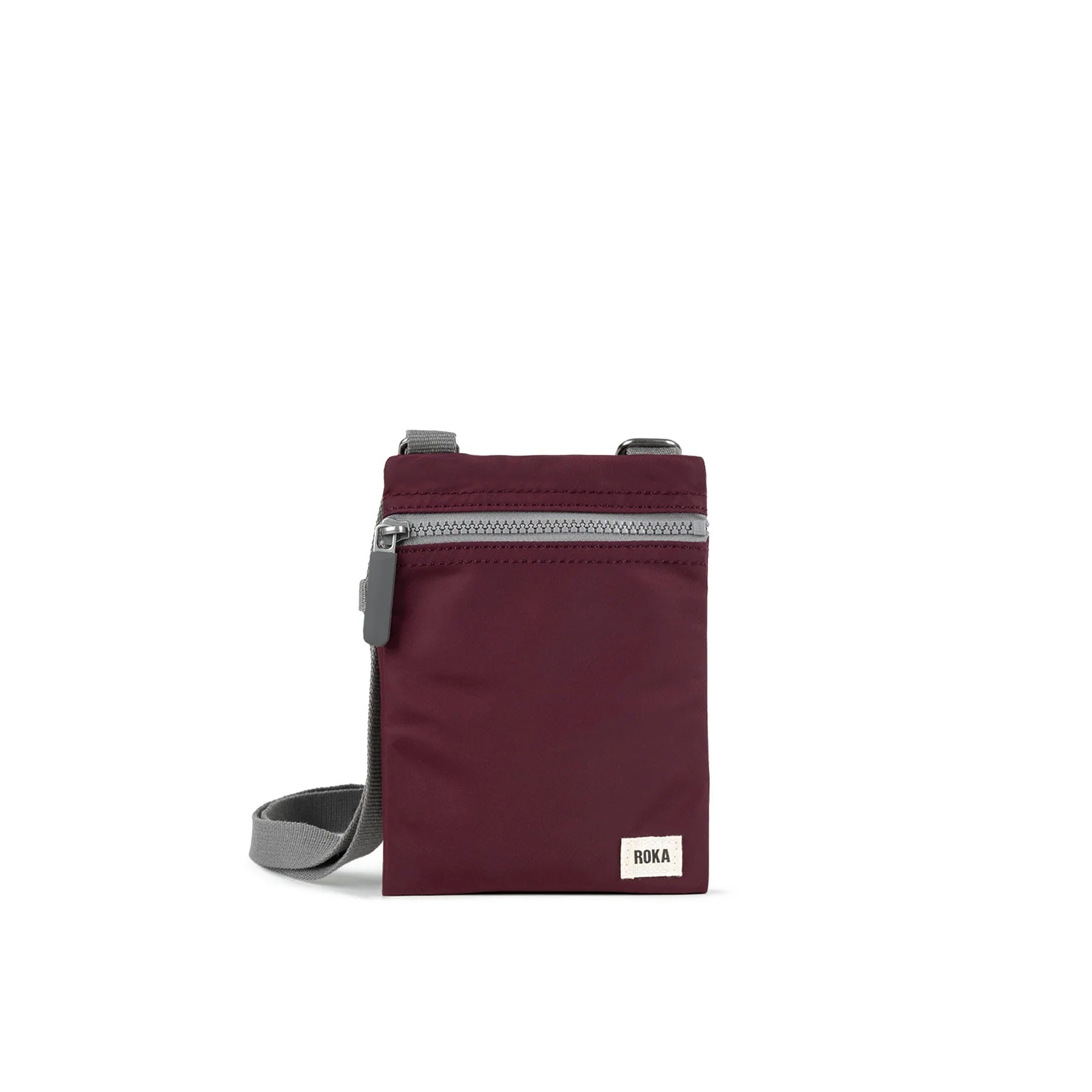 A photo of the front of a small rectangular dark plum coloured pocket bag. It has a grey zip horizontally at the top, grey straps, and a small ROKA logo in the bottom right corner.