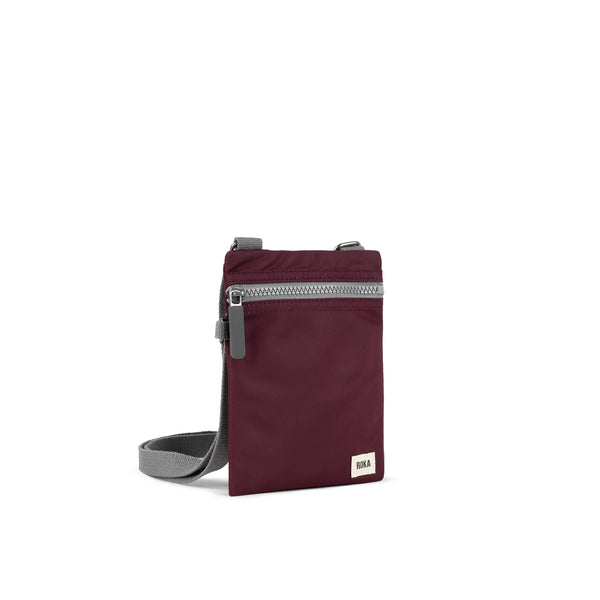 A photo of the front and side of a small rectangular dark plum coloured pocket bag. It has a grey zip horizontally at the top, grey straps, and a small ROKA logo in the bottom right corner.