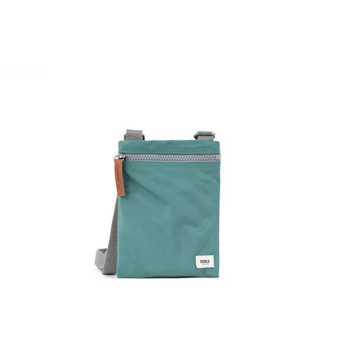 A photo of the front of a small rectangular sage green pocket bag. It has a grey zip horizontally at the top, grey straps, and a small ROKA logo in the bottom right corner.