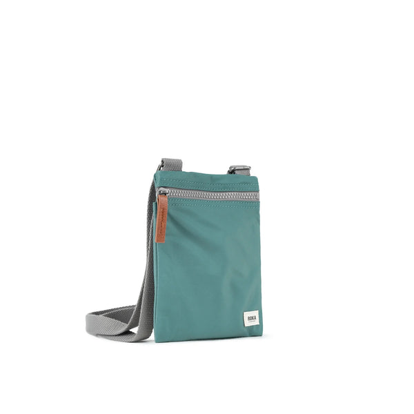  A photo of the front and side of a small rectangular sage green pocket bag. It has a grey zip horizontally at the top, grey straps, and a small ROKA logo in the bottom right corner. The zip pull is brown.