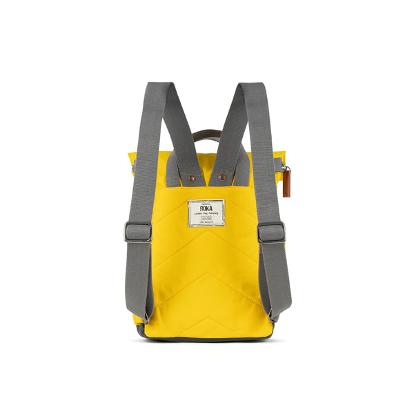 Photo showing the back of a small yellow coloured backpack. The straps and handles are grey and there’s a small fabric label with the ROKA logo on.