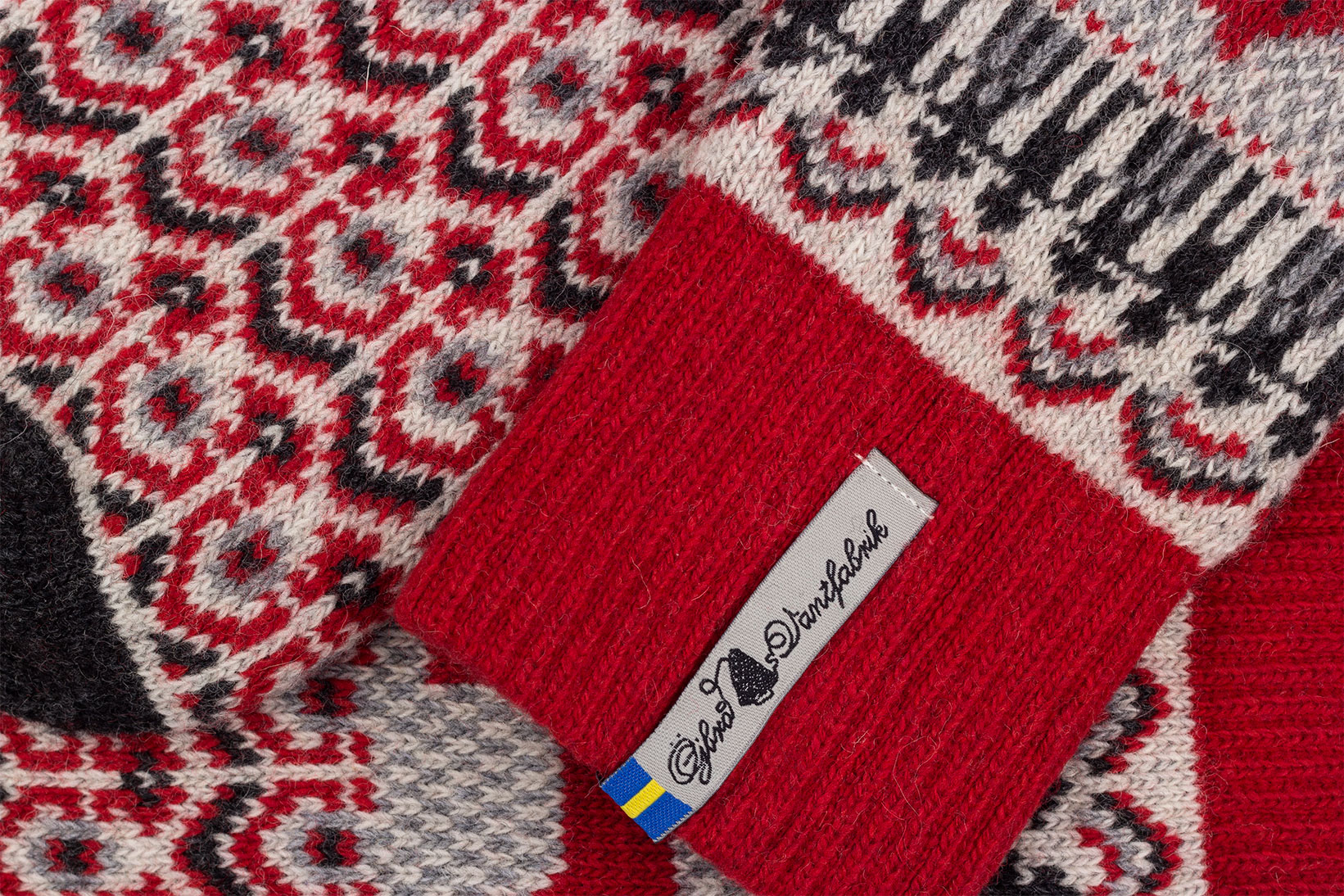 Close-up of a knitted socks showing a red ribbed cuff with a white tag alongside a red, gray, and black geometric pattern.